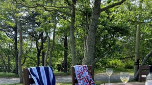 Eine Holzbank mit einer gestreiften Decke, ein Tisch mit einem Weinglas und einer Flasche, und Blick auf ein bewaldetes Gebiet.