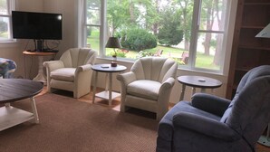 Ein Wohnzimmer mit drei Sesseln, einem Couchtisch und einem Fernseher auf einem Schrank. Große Fenster bieten Blick auf einen Garten.