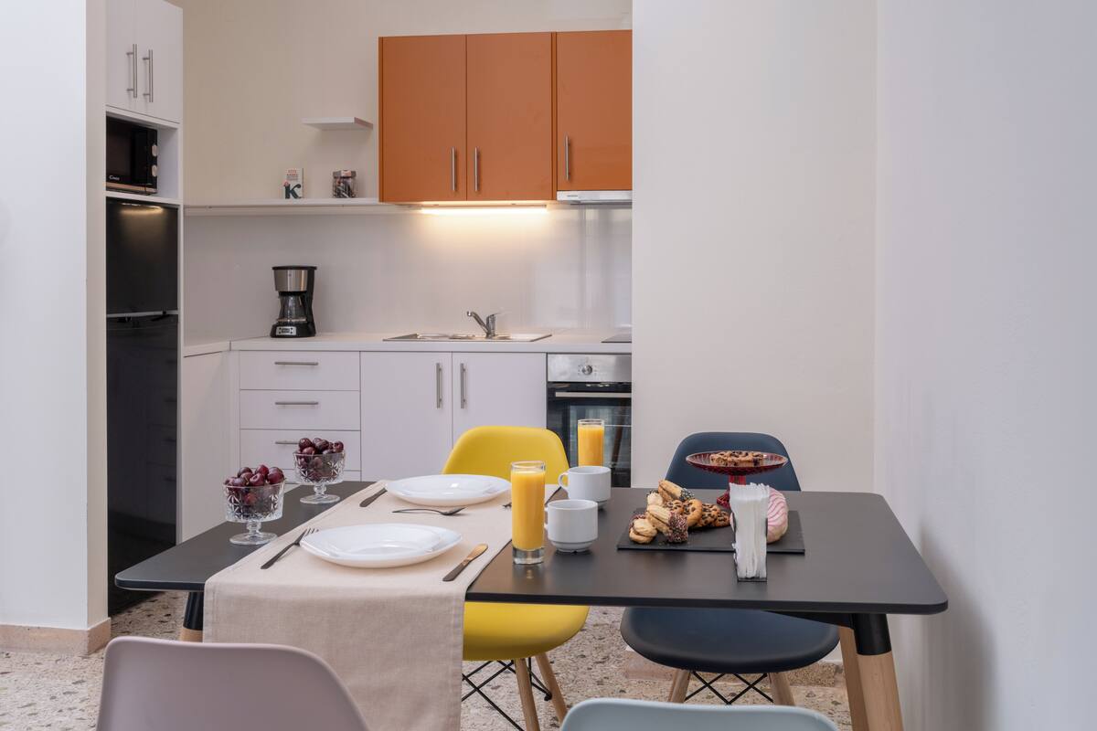 Een compacte keuken met witte en oranje kastjes, een koffiezetapparaat en een eettafel met borden, bestek en een glas sinaasappelsap.