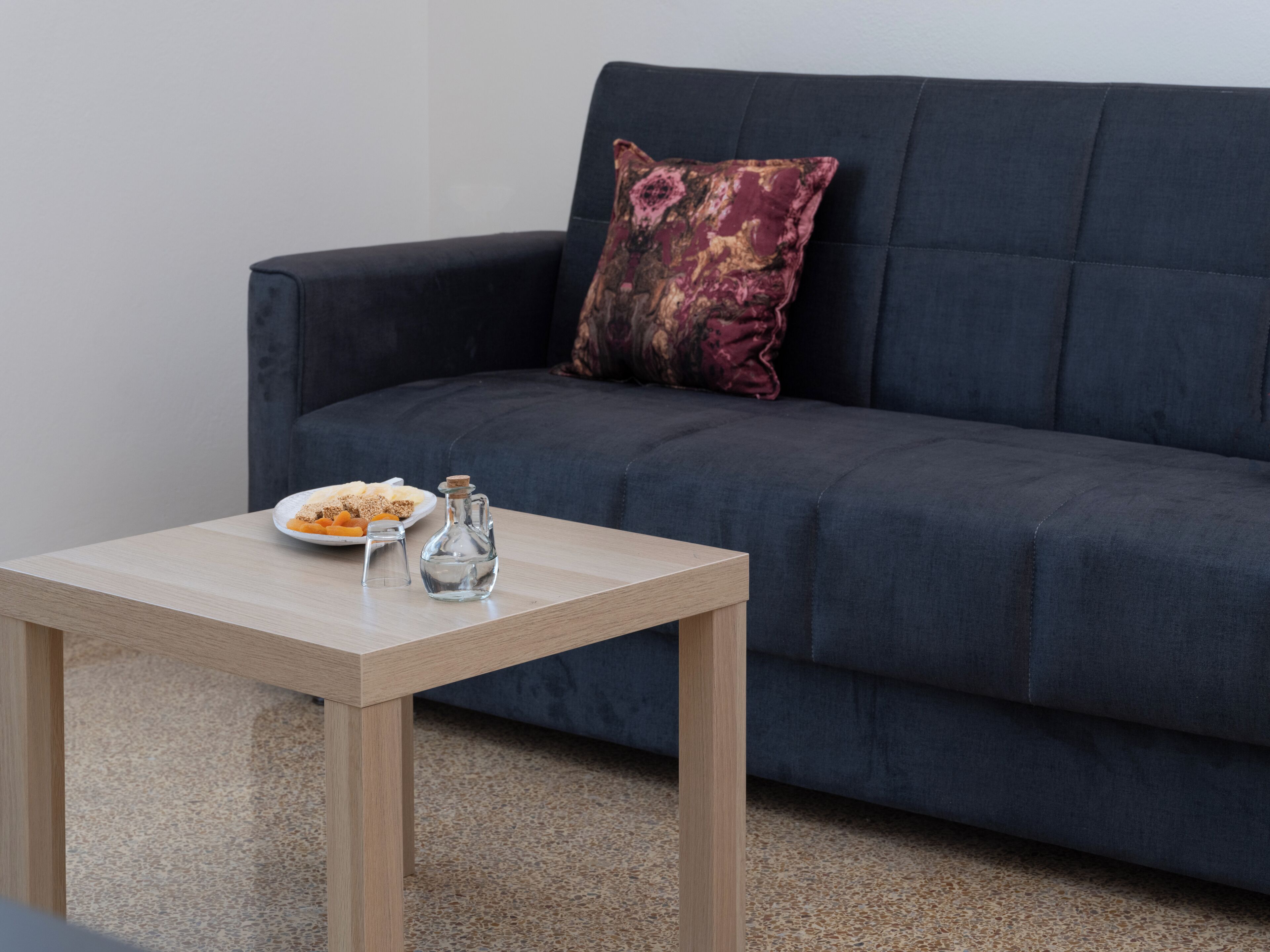 A modern living room with a dark blue sofa, a small wooden coffee table, a decorative pillow, and a plate of snacks.