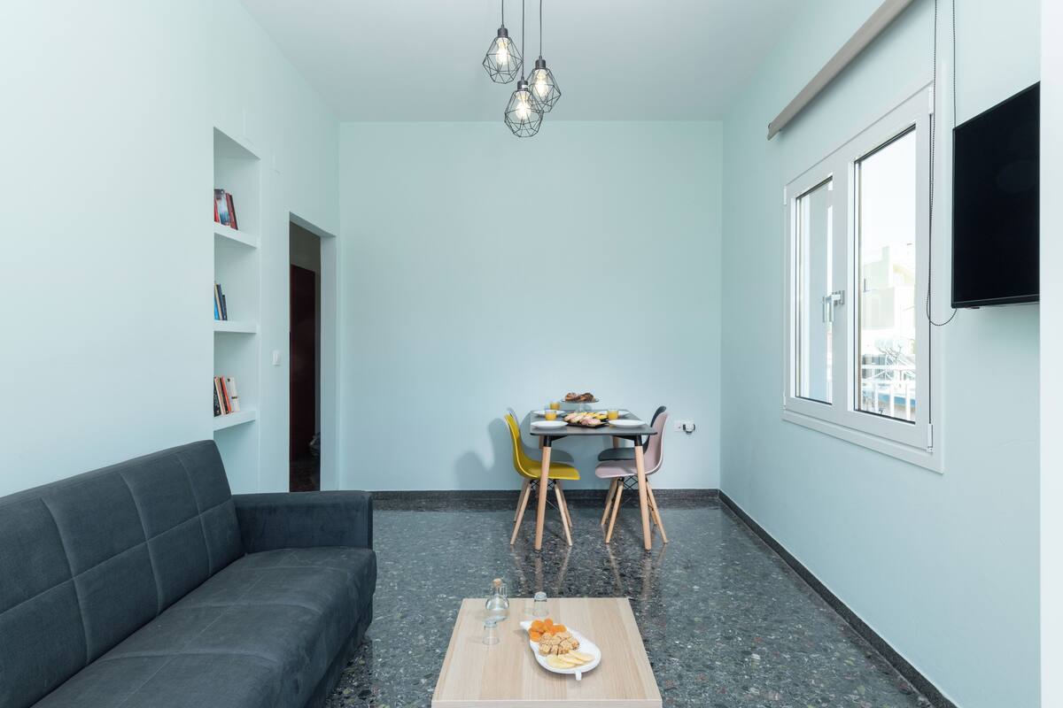 Een moderne woonkamer met een grijze bank, een eettafel met stoelen en een aan de muur bevestigde televisie.