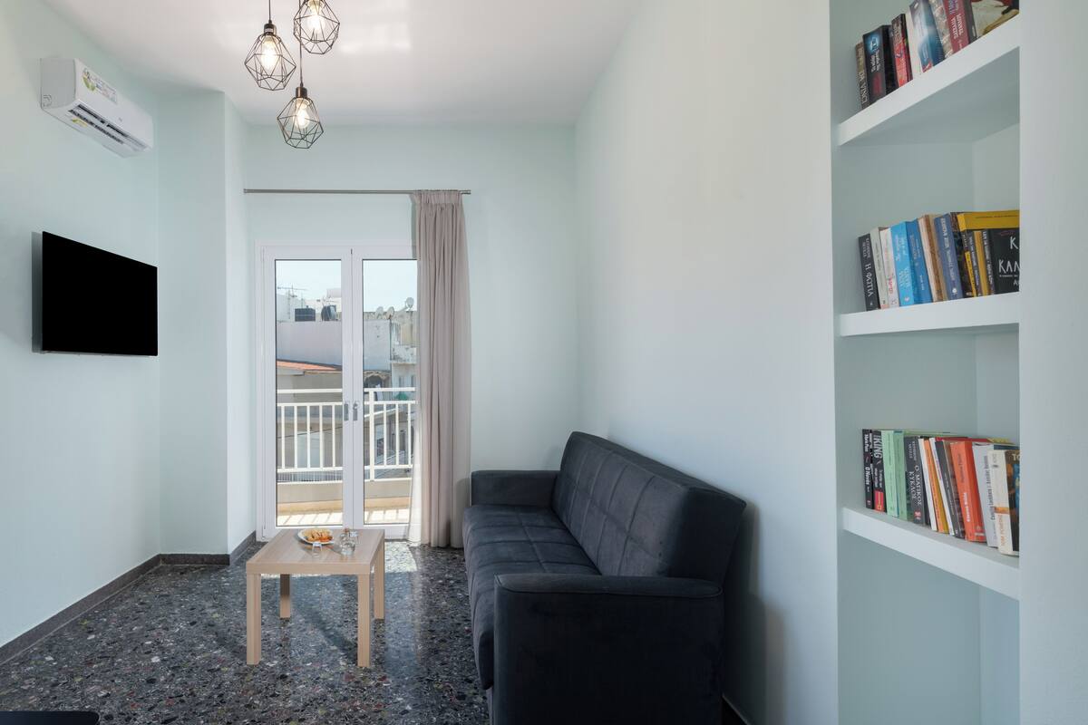 Een moderne woonkamer met een grijze bank, een klein houten tafeltje, een boekenkast en een aan de muur bevestigde televisie.