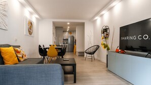 Moderni olohuone, jossa on sohva, ruokapöytä ja tuoleja. Seinälle on asennettu televisio, ja taustalla näkyy keittiö.