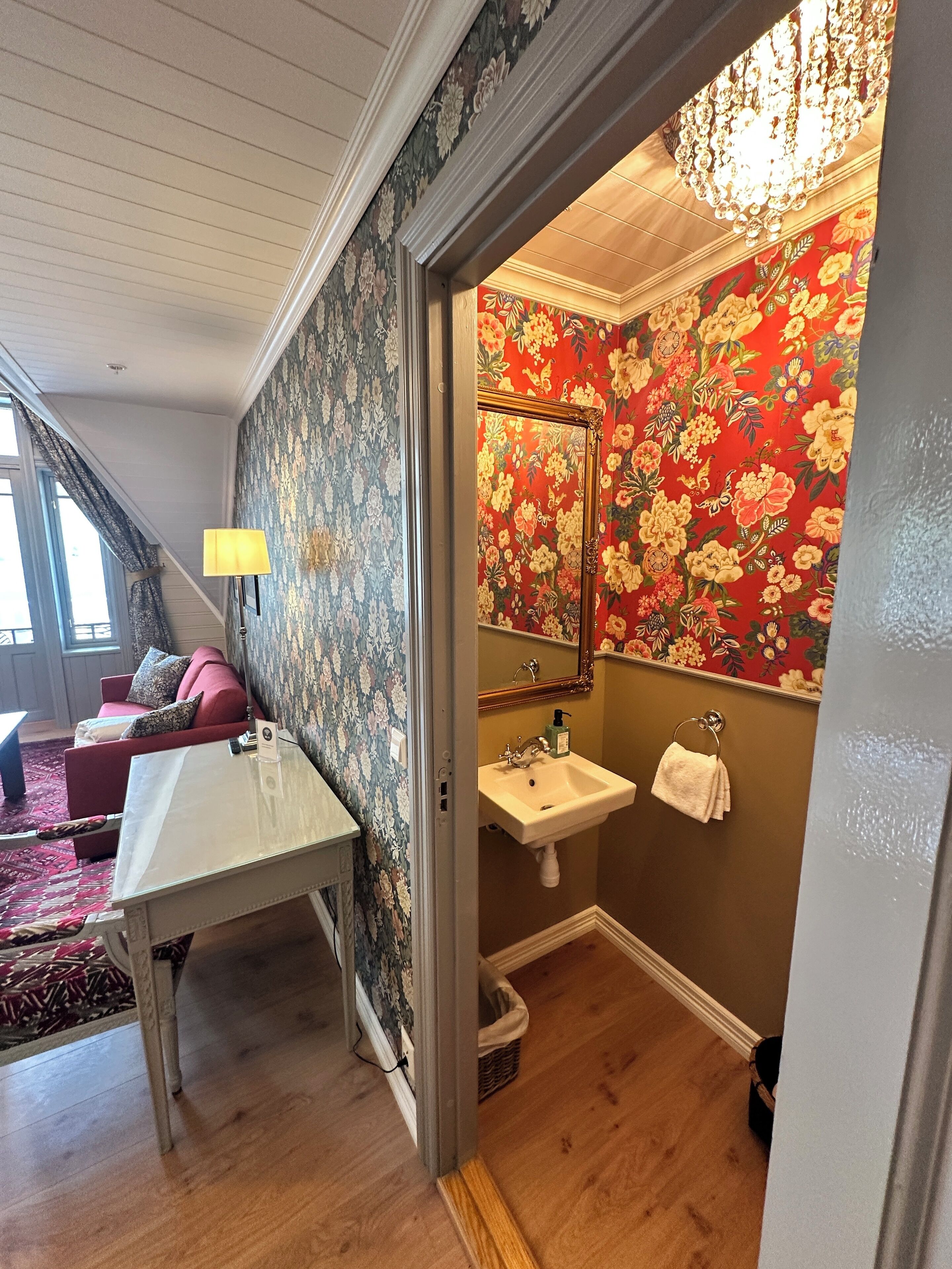 A room with a wooden floor, a red sofa, a white table, a chandelier, a sink, and a mirror.