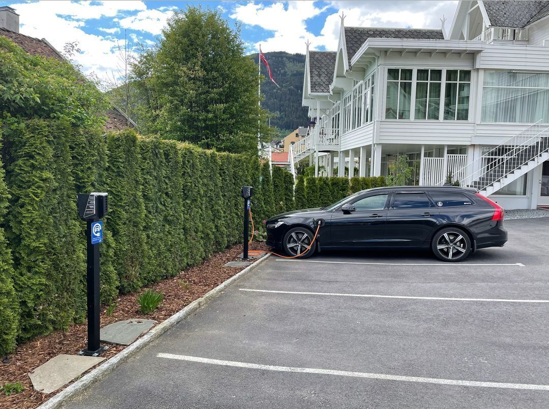 A black car parked in a driveway with a charging station, a hedge, and a building with a balcony in the background.