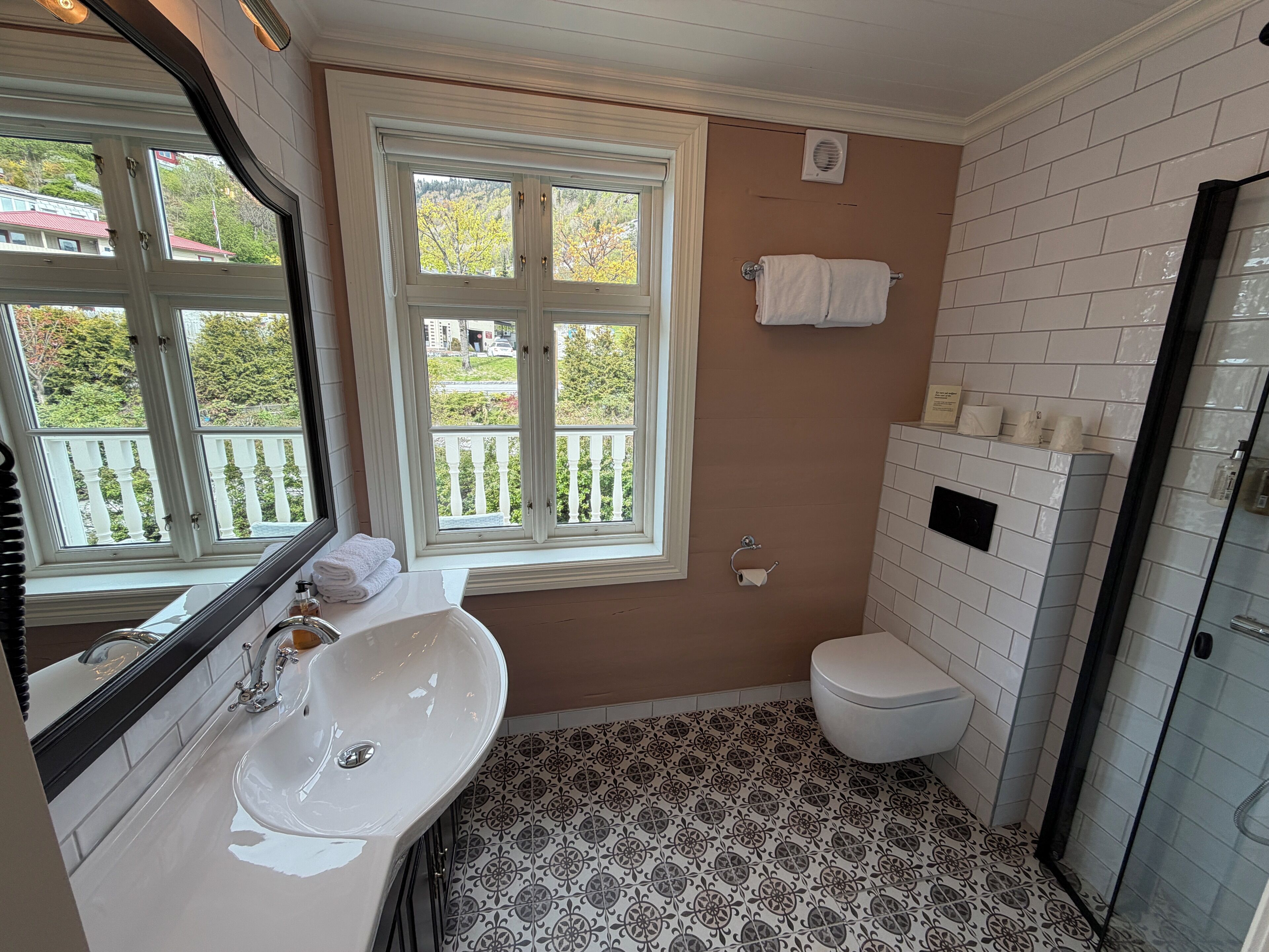 Un baño con lavamanos blanco, inodoro, ventana con vista a follaje y zona de ducha.