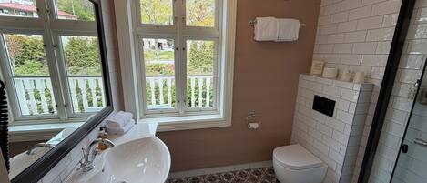 Un baño con lavamanos blanco, inodoro, ventana con vista a follaje y zona de ducha.