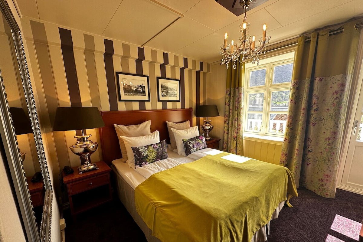 Habitación de hotel con una cama grande, dos mesitas de noche, un candelabro, una ventana con cortinas y un espejo.