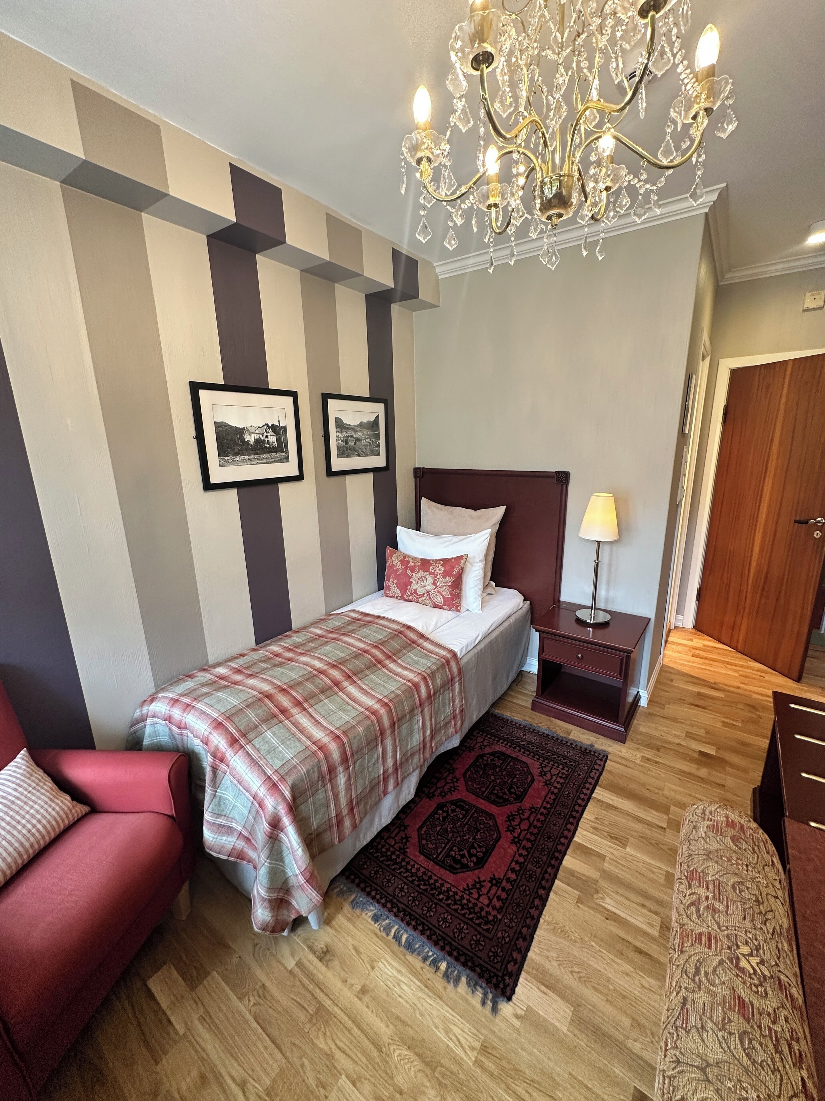 A bedroom with a bed, a red armchair, a nightstand, a lamp, a chandelier, and two framed pictures on the wall.
