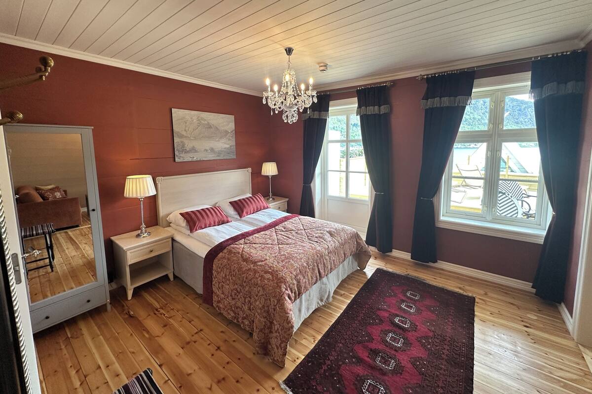 Un dormitorio con una cama grande, una mesita de noche, un aparador, un espejo, una araña y vistas al exterior a través de una ventana.