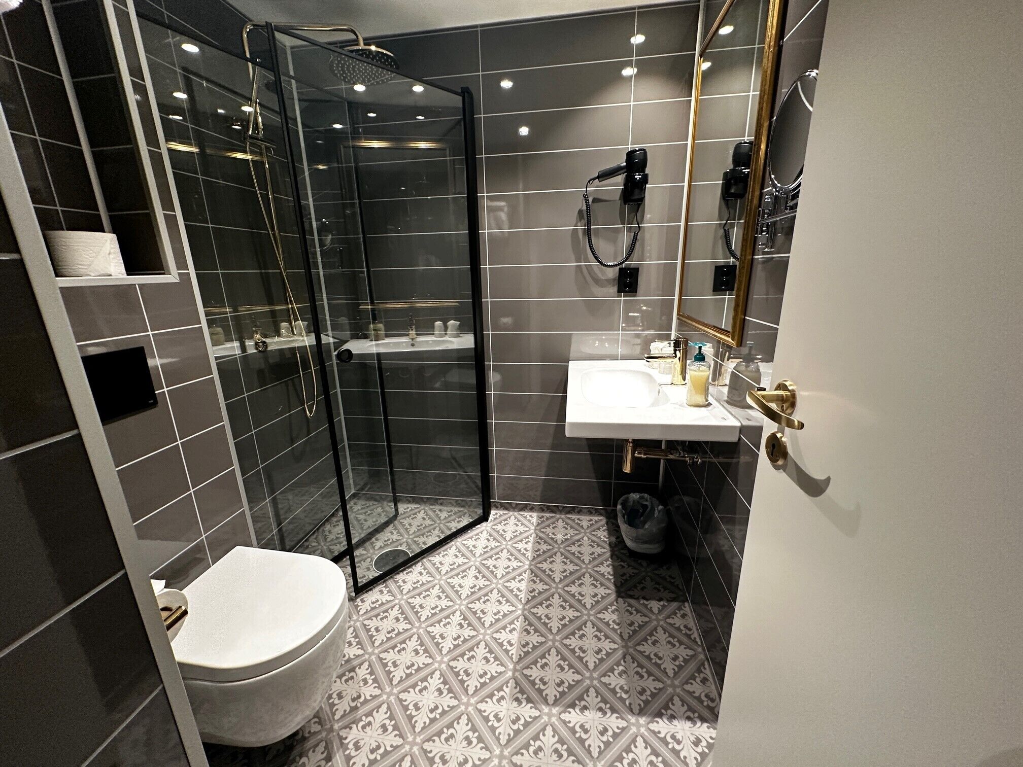 A modern bathroom with a glass-enclosed shower, a white sink with gold fixtures, and patterned floor tiles.