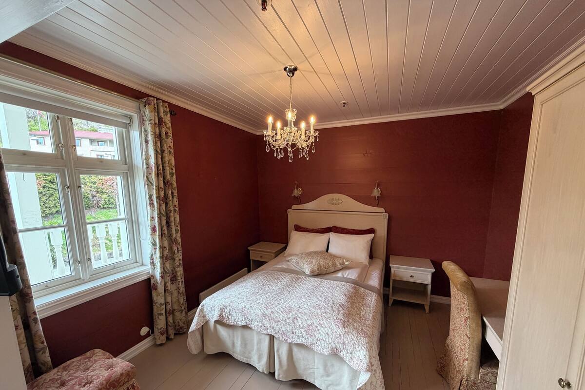 Un dormitorio con cama, mesitas de noche, una araña, una ventana con cortinas y una puerta.