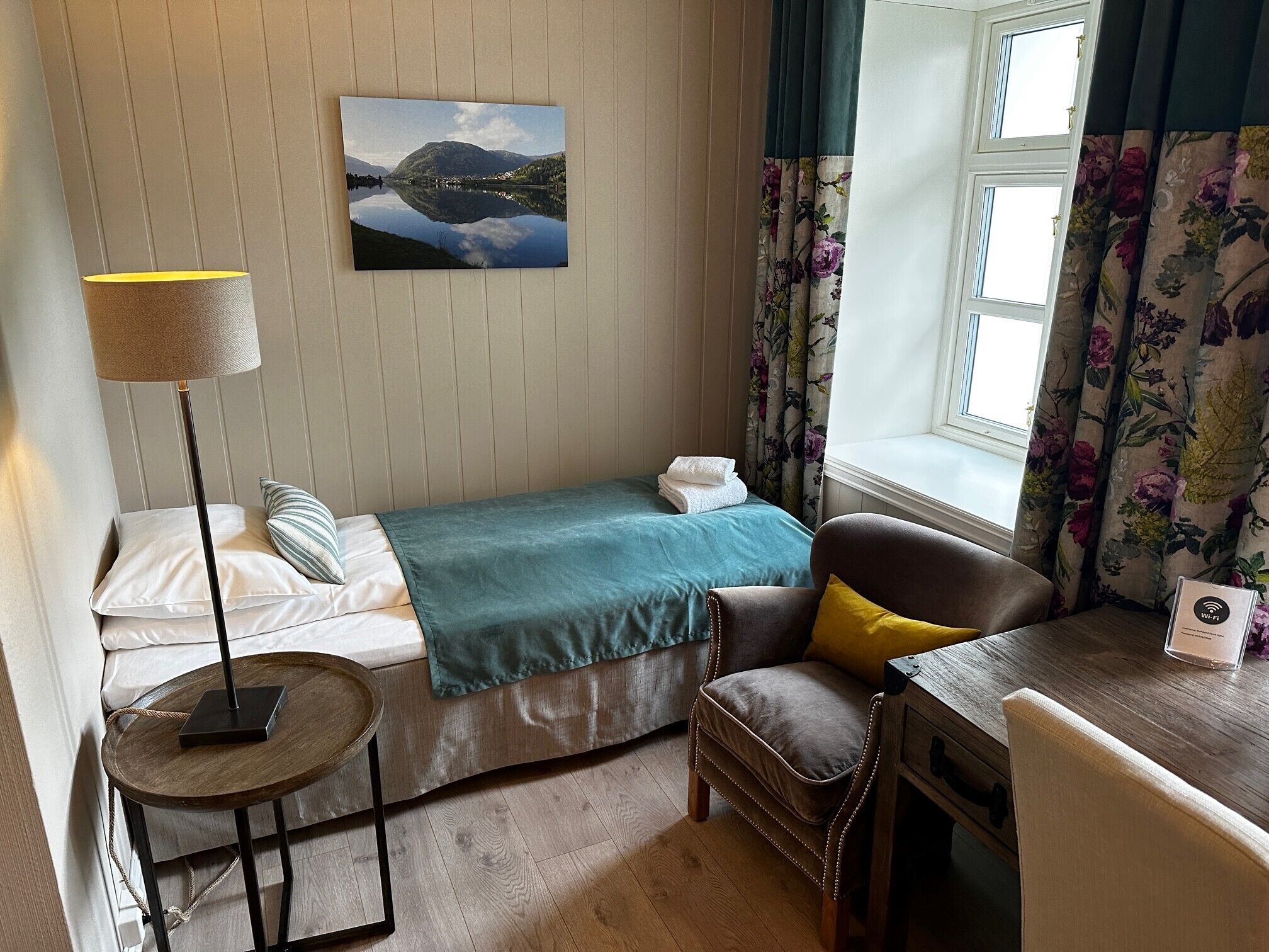 A neatly arranged bedroom with a bed, a chair, a small table, a lamp, a painting, and a window with curtains.