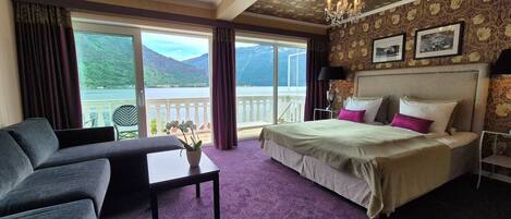 A hotel room with a bed, a sofa, a table, and a view of a lake and mountains.