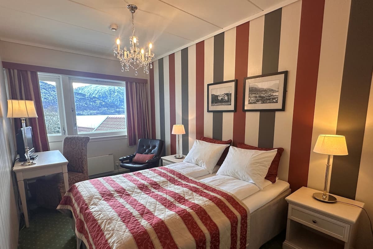 Una habitación de hotel con cama, escritorio, sillón, candelabro y ventana con cortinas.