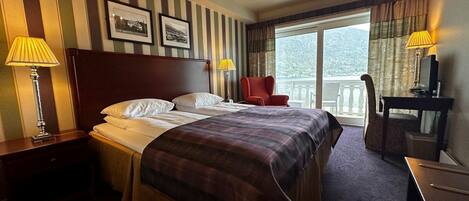 A hotel room with a large bed, a desk with a computer, a chair, a nightstand, a lamp, a chandelier, and a balcony with a view.