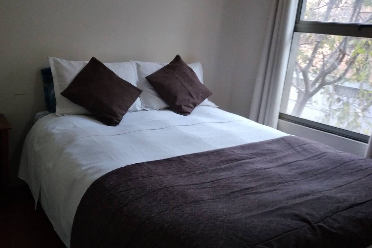 A hotel room with a bed, pillows, a nightstand, and a window.