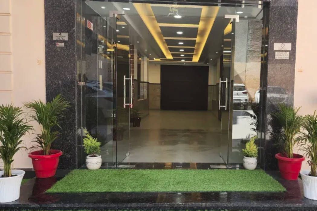 Entrada de hotel con puerta de vidrio, felpudo de césped artificial verde y plantas en macetas a ambos lados.