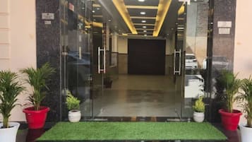 Entrada de hotel con puerta de vidrio, felpudo de césped artificial verde y plantas en macetas a ambos lados.