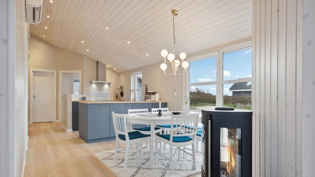 A modern kitchen with a central island, a dining area with white chairs and a blue tablecloth, and a large window offering a view of the outdoors. - Beautiful home in Tarm with WiFi (Tarm)