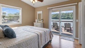 Quarto com cama, ventilador de teto, cômoda, espelho e porta de correr para uma varanda com vista para um lago e árvores.