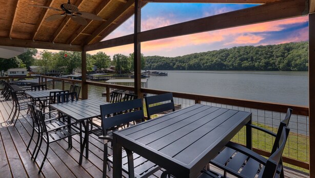Um deck externo com mesas e cadeiras, com vista para um lago e colinas arborizadas.