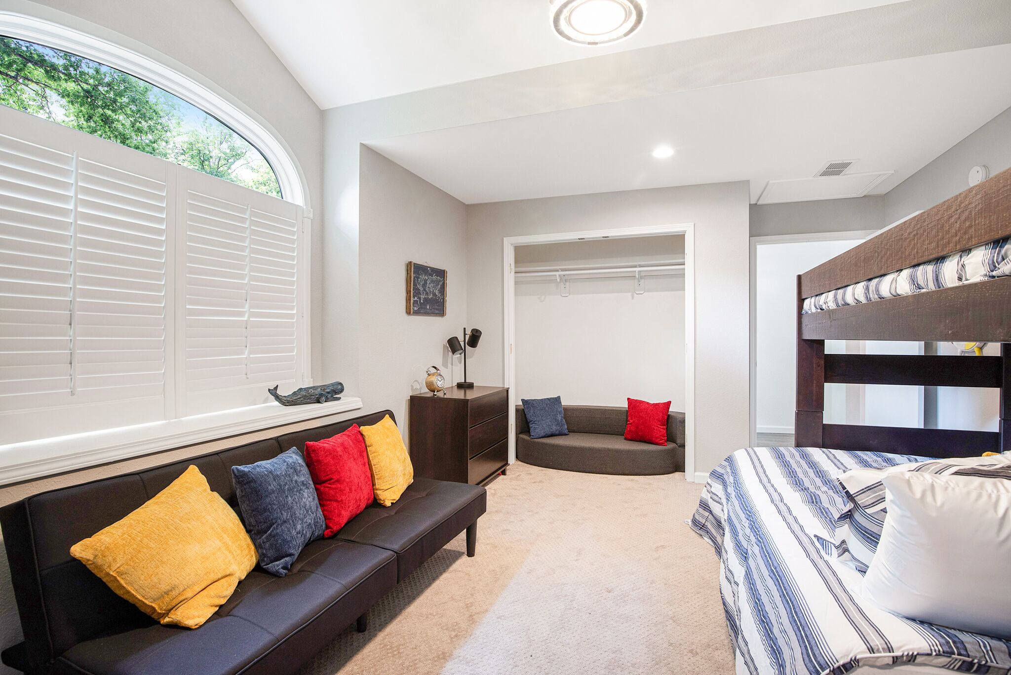 A bedroom with a bunk bed, a sofa with colorful cushions, a bedside table, and a large window with blinds.