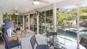 Une pièce lumineuse, baignée de soleil, avec une grande fenêtre donnant sur une étendue d’eau, et meublée d’un canapé, de fauteuils et d’une table en verre.