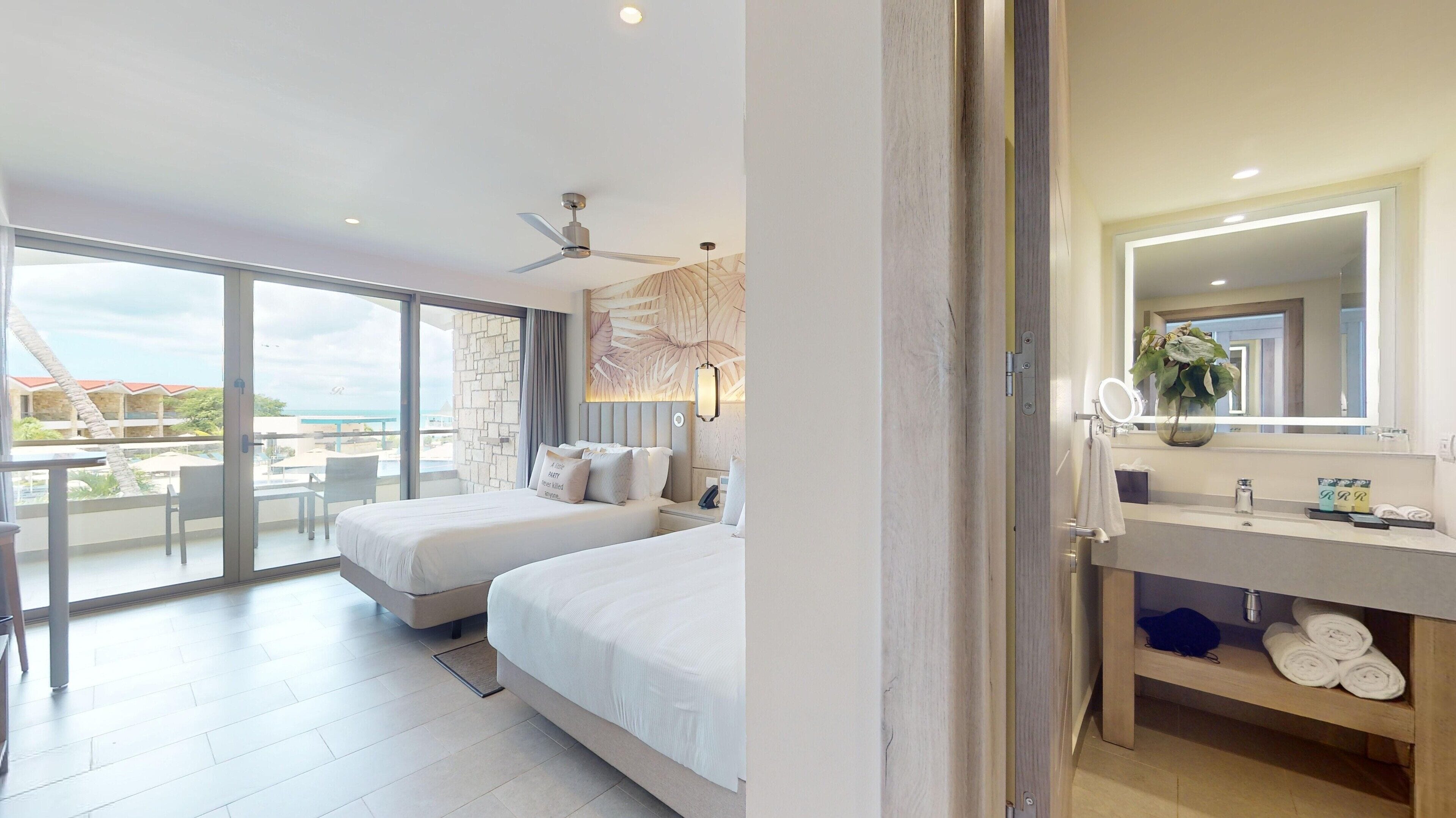 A hotel room with two beds, a balcony with a view, a modern bathroom with a large mirror, and a ceiling fan.