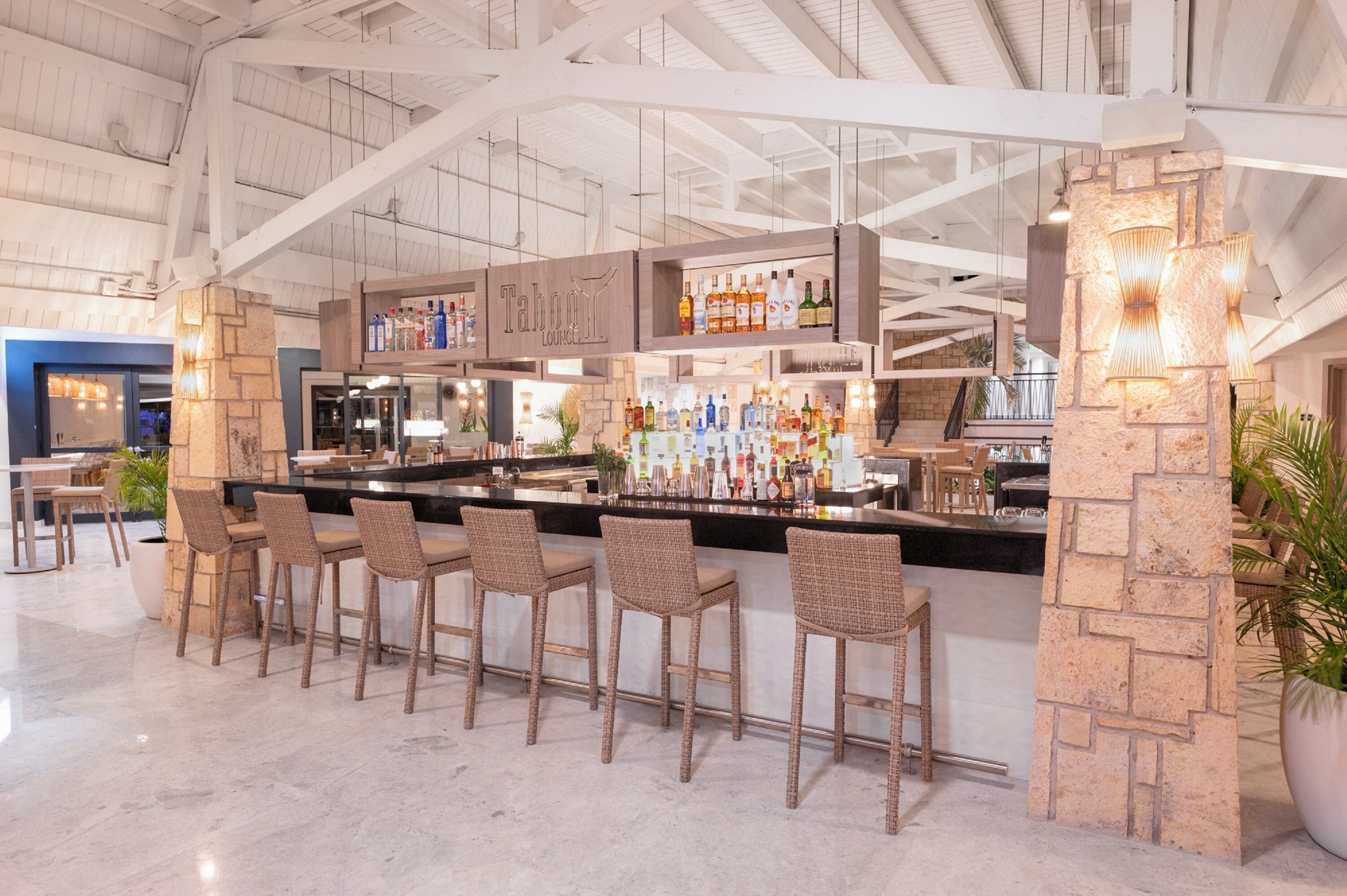 A bar area with a stone pillar, wicker bar stools, and a variety of bottles on shelves.