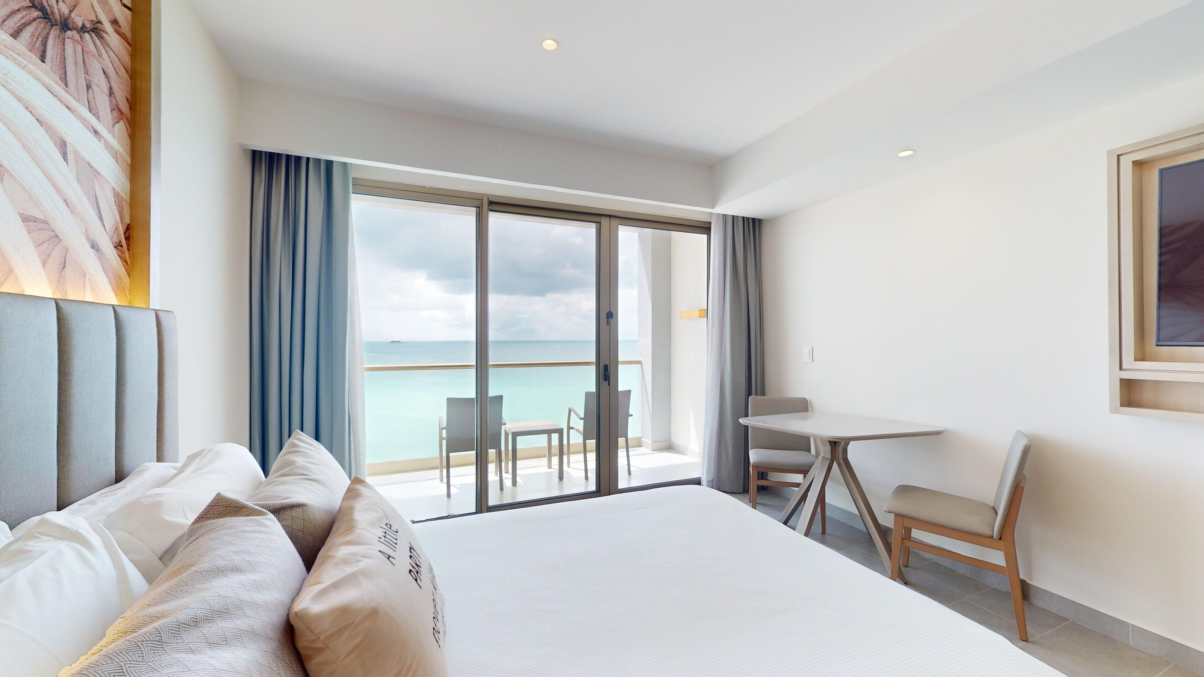 A hotel room with a large bed, a small table, and a chair. There is a balcony with a view of the ocean.