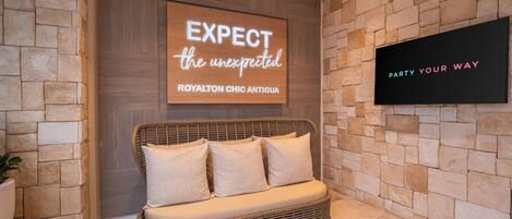 A lobby with a stone wall, a wicker sofa with cushions, a wooden sign with text, and a TV screen.