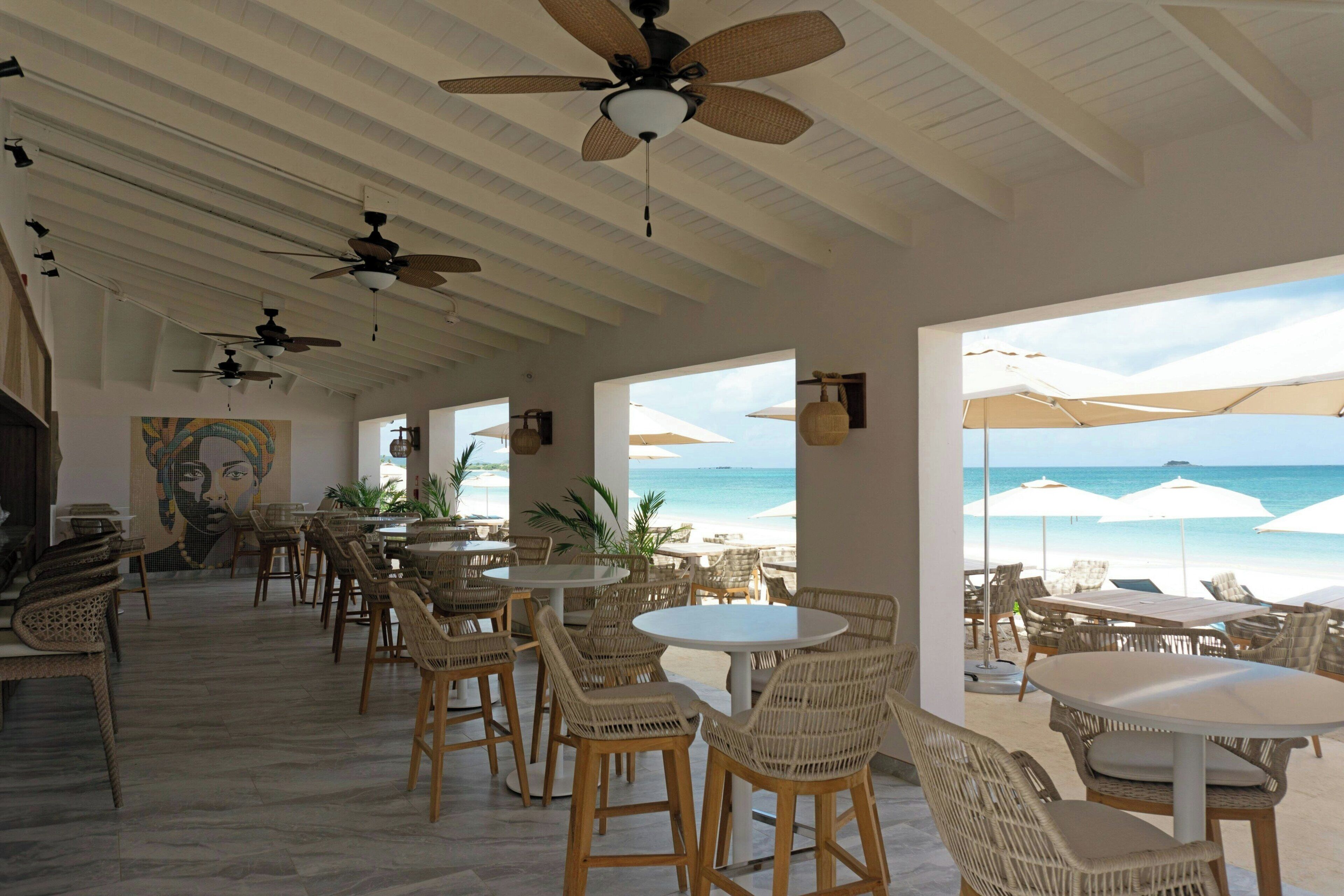 Un restaurant situé en bord de mer, doté de meubles en osier, de ventilateurs de plafond et de grandes fenêtres donnant sur l’océan.