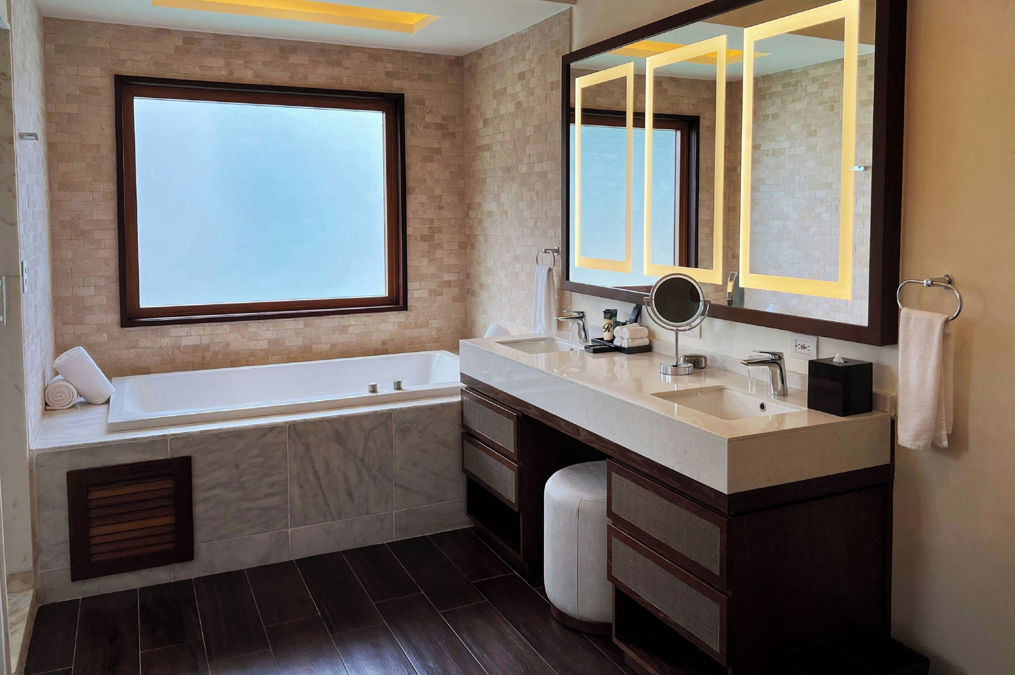 Un baño moderno con una bañera grande, dos lavamanos, un tocador con cajones, un espejo amplio y una ventana que proporciona luz natural.