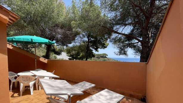 A patio with white tables and chairs, a green umbrella, and a view of trees and the sea.