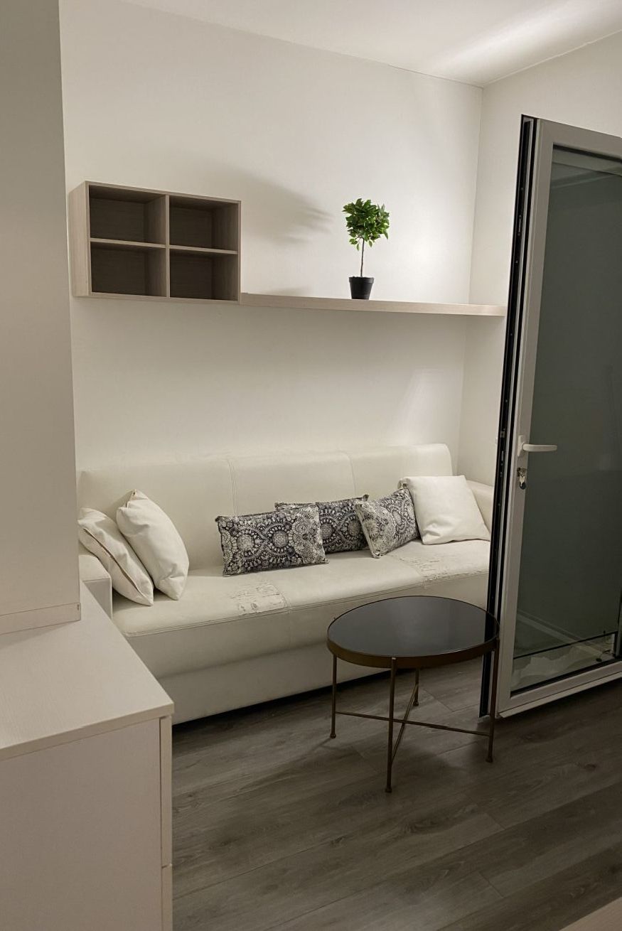 A modern living room with a white sofa, patterned cushions, a small round table, a potted plant, and a glass door.