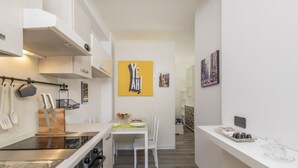 A narrow kitchen with white cabinetry, a central island with a sink, and a dining area with chairs and a table. - ARCHIMEDE, 53 · Apt 3 - Monolocale con spazio esterno (Milano)