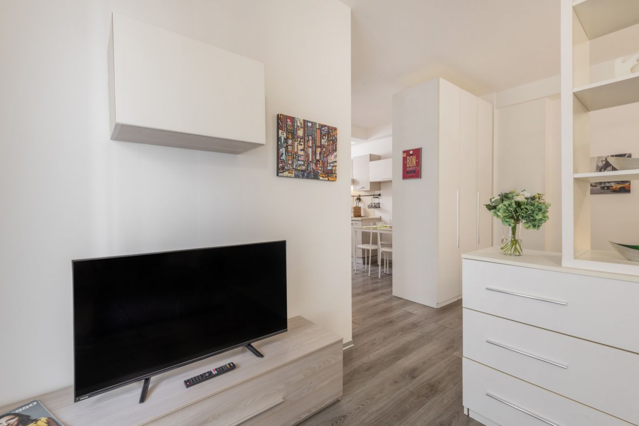 A modern living room with a flat-screen TV on a wooden stand, a white cabinet with drawers, a small table with a vase of flowers, and a colorful abstract painting on the wall.