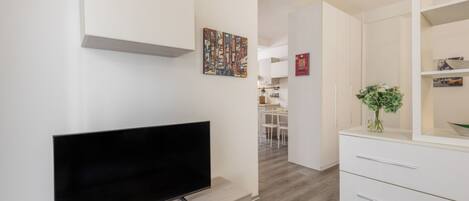 A modern living room with a flat-screen TV on a wooden stand, a white cabinet with drawers, a small table with a vase of flowers, and a colorful abstract painting on the wall.