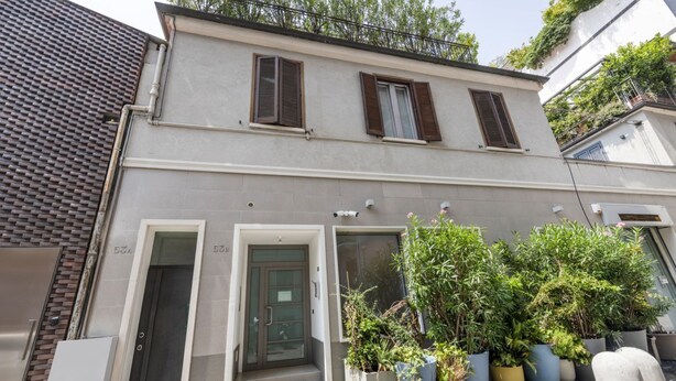 A two-story building with a grey facade, two windows with brown shutters, and a door. There are potted plants in front of the building. - ARCHIMEDE, 53 · Apt 3 - Monolocale con spazio esterno (Milano)