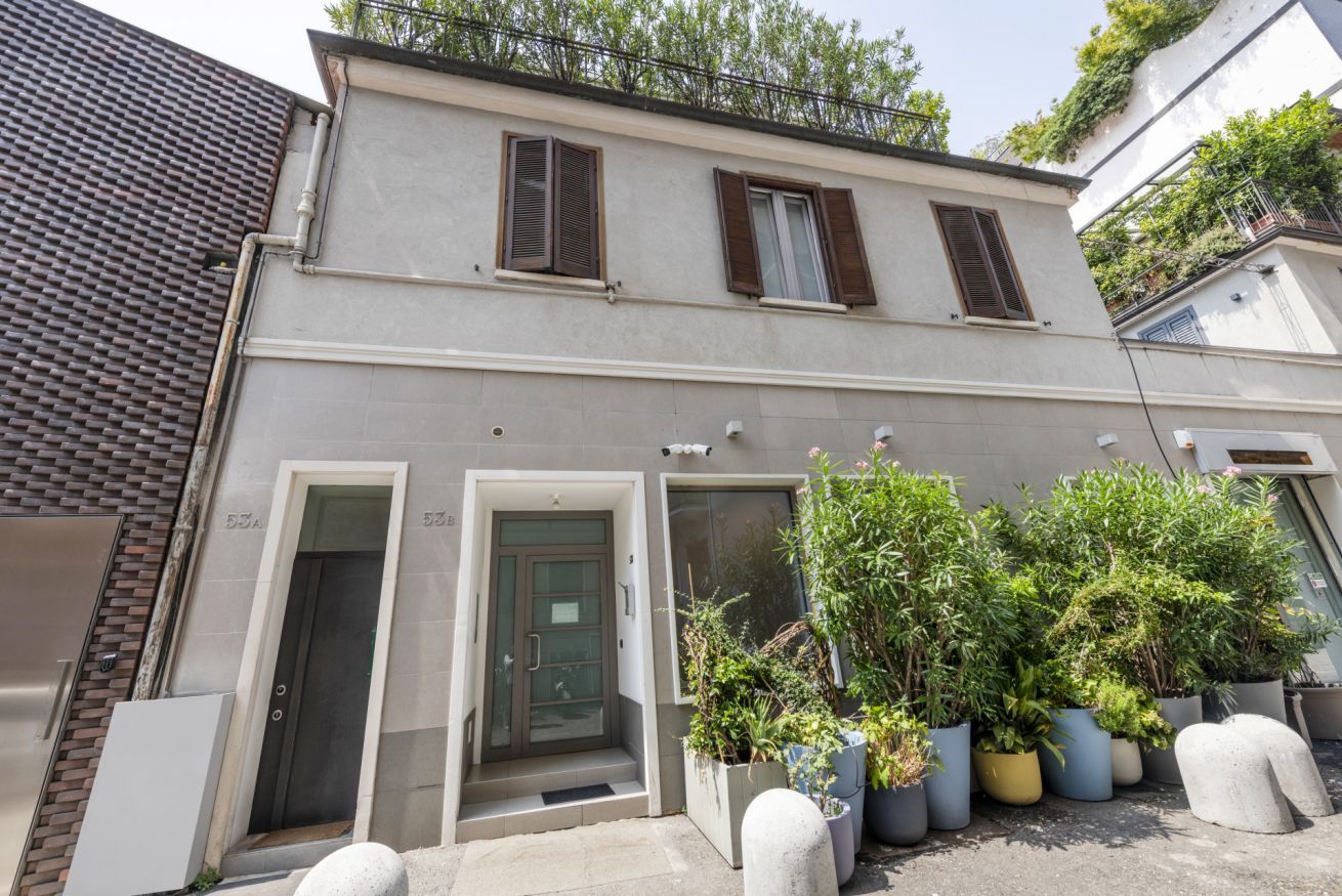 Un immeuble de deux étages avec une façade grise, deux fenêtres dotées de volets bruns et une porte. Des plantes en pot sont disposées devant l’immeuble.