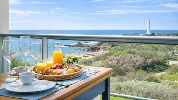 A balcony with breakfast items, a view of the ocean, and a lighthouse in the distance.