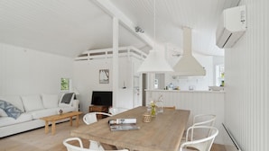 Amplia sala de estar con una mesa de comedor de madera, sillas blancas, un sofá blanco y un televisor.