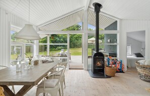 Een ruime kamer met een grote houten tafel, witte stoelen, een open haard en grote ramen met uitzicht op de omgeving.