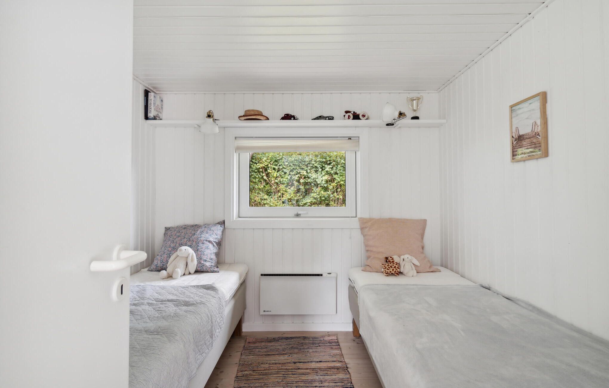 Una habitación pequeña y blanca con dos camas individuales, una ventana que da a zonas verdes, un radiador y una estantería con diversos objetos.