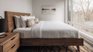 Une chambre avec un lit en bois, du linge de lit blanc, une table de chevet, un tableau accroché au mur et une grande fenêtre donnant sur des arbres.