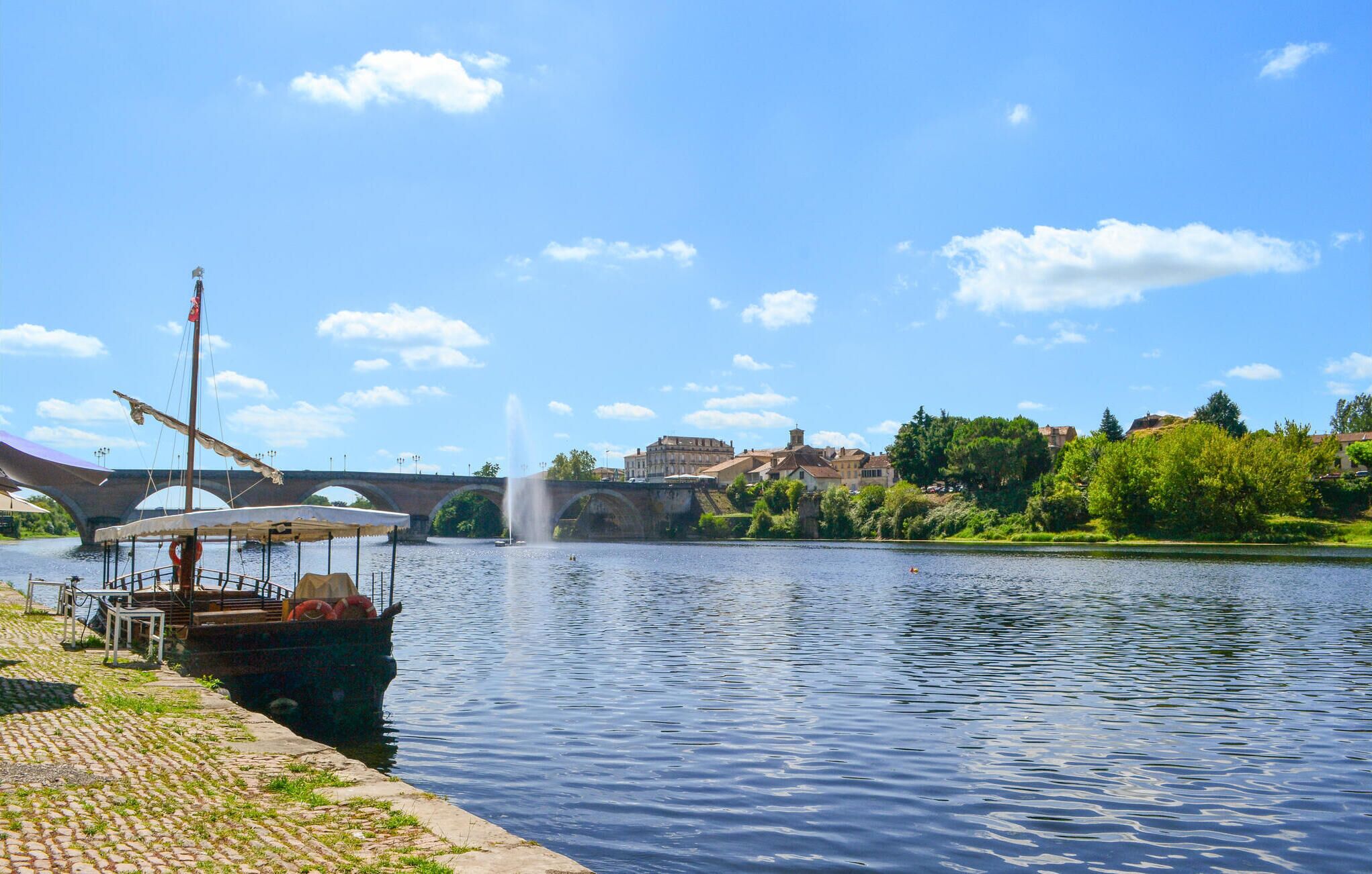 A river with a boat docked on the left, a bridge in the background, and buildings on the opposite bank.