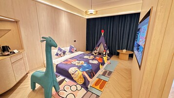Habitación de hotel con cama que tiene una colcha temática de Mickey Mouse, una mesita de noche con forma de jirafa, una estructura tipo tienda de campaña y un televisor en la pared.