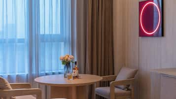A hotel room with a round table, two chairs, a vase with flowers, a bottle, and a neon sign on the wall.