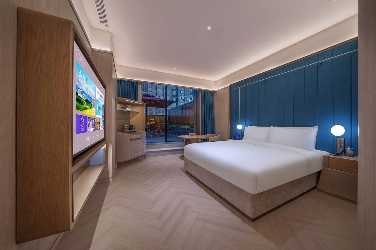 A modern hotel room with a large bed, a TV on a stand, a desk, and a view of the outdoors.